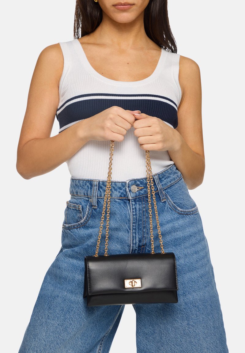 Borsa a tracolla in pelle nera con chiusura dorata e tracolla a catena, forma rettangolare, texture liscia, tenuta da una persona che indossa una maglietta bianca e jeans blu.