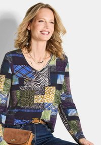 Langärmliges Oberteil mit einem Patchwork-Design in Grün, Blau, Gold und Schwarz, welches Tierdrucke und Karomuster beinhaltet, kombiniert mit Jeans.