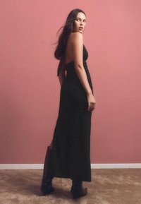 Fekete, ujjatlan maxi ruha, testhez simuló sziluettel, oldalsó hasítékkal és nyitott hátrészen, magas fekete csizmával viselve, rózsaszín háttér előtt.
