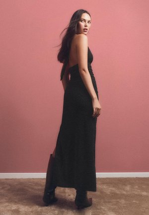 Fekete, ujjatlan maxi ruha, testhez simuló sziluettel, oldalsó hasítékkal és nyitott hátrészen, magas fekete csizmával viselve, rózsaszín háttér előtt.