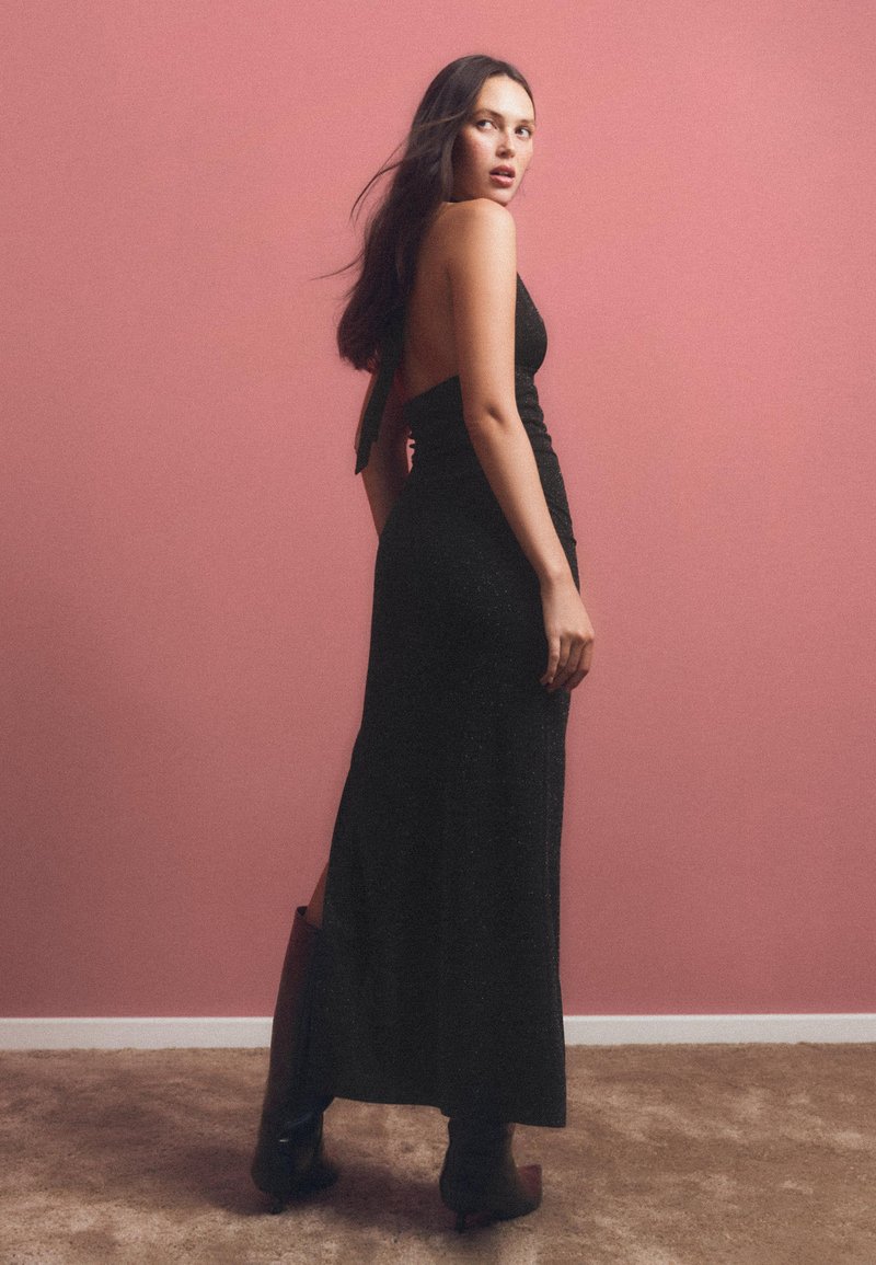 Rochie maxi neagră, fără mâneci, cu o siluetă ajustată, fante pe lateral și spate descoperit, purtată cu cizme negre înalte pe un fundal roz.