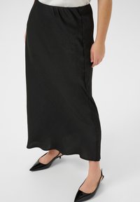 Jupe maxi en satin noir avec une texture lisse, arborant un motif subtil, une taille élastique et une silhouette fluide, accompagnée de chaussures noires à bout pointu.