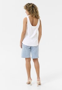 Weißes ärmelloses Tanktop mit tiefem Rückenausschnitt, kombiniert mit hellblauen Jeansshorts und beigen Absatzsandalen, von hinten gesehen.
