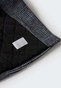 Doublure en tissu noir matelassé avec une couche extérieure à motif à carreaux gris, comportant une étiquette blanche pour le nom et les coordonnées téléphoniques sur le côté intérieur.