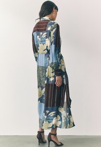 Fließendes Kleid mit einer Mischung aus floralen und gestreiften Mustern in Blau, Braun und Grün, mit langen Ärmeln und einem seitlichen Schlitz.