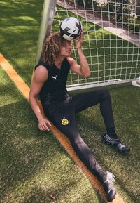 Atleet in een zwart mouwloos shirt en broek met een geel logo, zittend op het gras met een voetbal op zijn voorhoofd, naast een doelpaal.