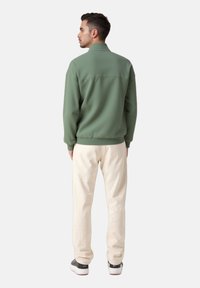 Veste synthétique verte à col montant et manches longues, associée à un pantalon couleur crème et des baskets noires. Texture lisse.