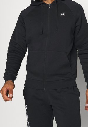 Mężczyzna ubrany w czarną bluzę z kapturem i suwakiem oraz dopasowane czarne spodnie dresowe z logo Under Armour na obu ubraniach.