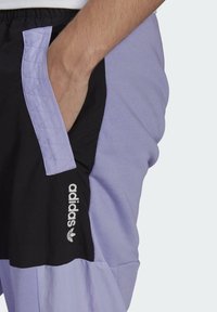 En persons hand i fickan på svarta och lavendelfärgade Adidas-shorts med logotyp på vänster lår, mot en enfärgad bakgrund.