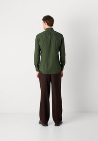 Cămașă lungă cu mâneci, de un verde închis, cu guler ascuțit, croială ajustată și manșete cu nasturi, asociată cu pantaloni largi maro și pantofi negri.