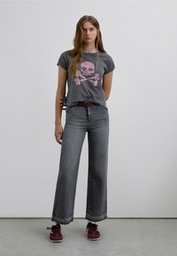 Camiseta gris con estampado de calavera y huesos cruzados en rosa, combinada con vaqueros anchos grises con sutiles adornos y zapatillas burdeos.