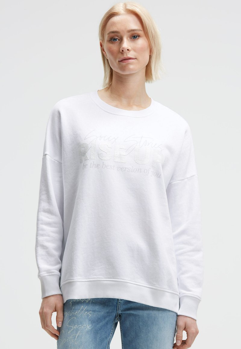 Blonde Frau in einem weißen Sweatshirt mit dem Text "Rise Up" und hellblauen Jeans, stehend vor einem einfarbigen hellgrauen Hintergrund.