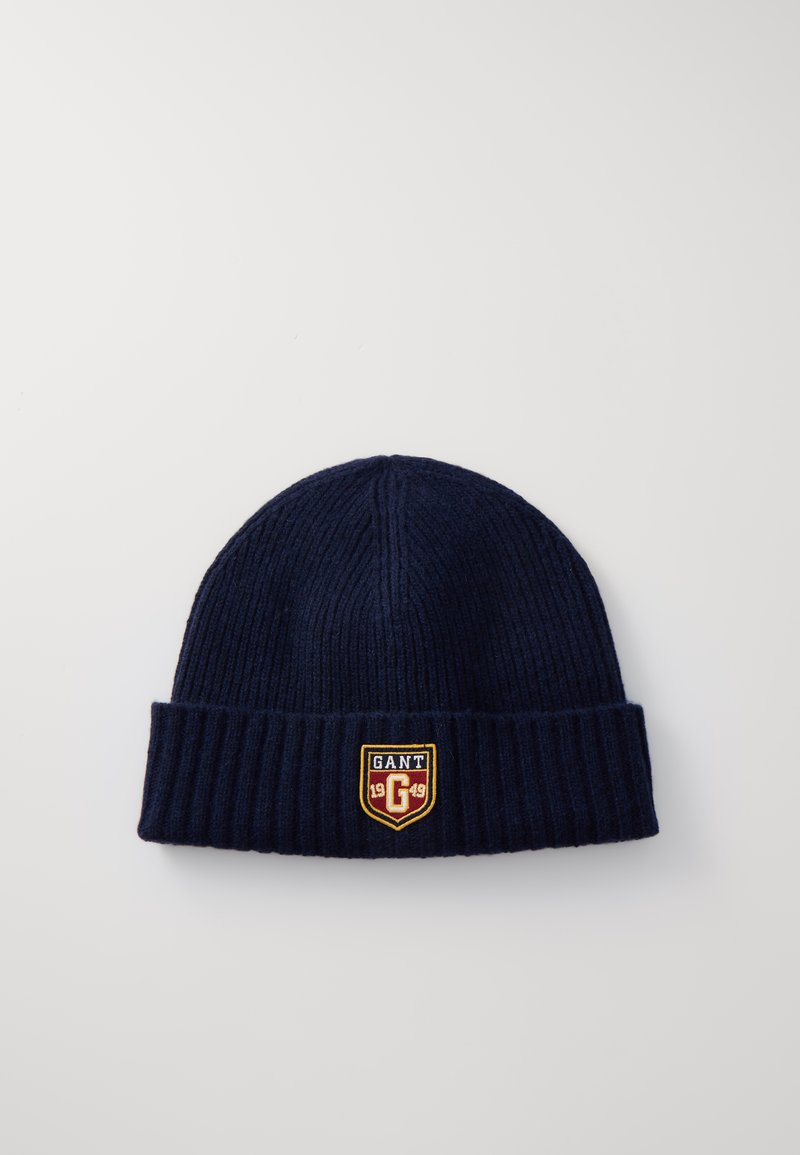 Navy blue knitted beanie made of ribbed fabric, featuring a folded cuff and a shield-shaped patch with "GANT" and "1949" in contrasting colors.