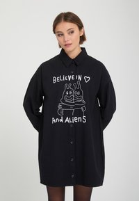 Jeune femme portant une robe chemise noire à boutons avec un graphisme blanc et le texte ""Believe in and aliens"" mettant en scène deux extraterrestres qui s'embrassent.