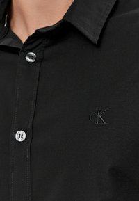 Camicia nera con colletto realizzata in tessuto liscio, caratterizzata da un sottile logo 'CK' ricamato e bottoni argentati lungo la patta.
