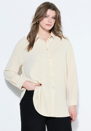 Junge Frau mit lockerem, welligem Haar, die ein cremefarbenes Hemd mit Knöpfen und schwarze Hose trägt und mit einer Hand in der Tasche posiert.