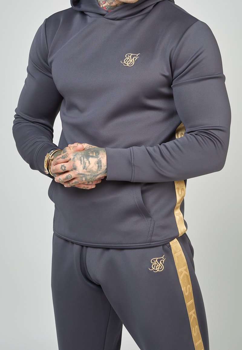 Homme aux mains tatouées portant un sweat à capuche gris foncé près du corps et un pantalon assorti avec un logo doré et une bande dorée sur le côté.