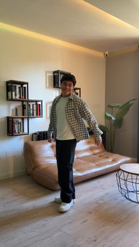 Una persona se encuentra en una habitación moderna, vistiendo una camisa de cuadros sobre una camiseta blanca, pantalones negros y zapatos blancos. Sofá de cuero marrón en el fondo.