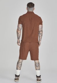 Braun gemustertes Polo- und Shorts-Set mit einem subtilen geometrischen Design; getragen mit dunklen Sneakern und weißen Sportsocken.