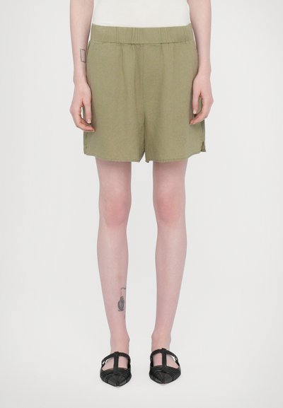 Shorts vert olive avec une taille élastique, en tissu léger, et des fentes sur les côtés. Associés à des chaussures noires à lanières.