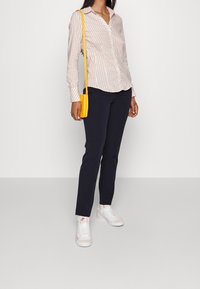 Camicia a righe a maniche lunghe in rosa chiaro e bianco, abbinata a pantaloni slim-fit blu navy e sneakers bianche. Una borsa a tracolla gialla aggiunge un tocco di colore.