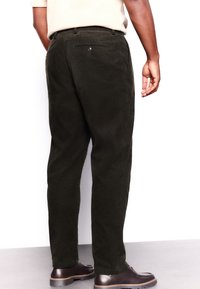 Pantalon en velours côtelé vert foncé à jambes droites, avec des poches arrière à boutons, porté avec des chaussures foncées. Tissu texturé visible.