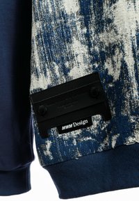 Suéter con tela texturizada en un patrón azul y blanco, mangas en contraste de color marino, y un parche de marca negro con acabado suave.