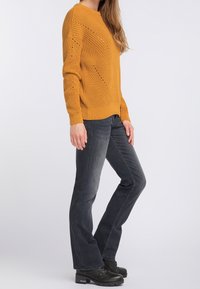 Nainen seisoo päällään sinapinkeltainen neule, tummanharmaat leveälahkeiset farkut ja mustat nauhakengät yksinkertaista vaaleaa taustaa vasten.