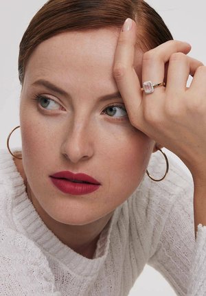 Kobieta z czerwonymi ustami i zielonymi oczami, nosząca złote kolczyki w kształcie kół oraz białą swetrową bluzkę, pokazuje złoty pierścionek z dużym prostokątnym przezroczystym kamieniem.