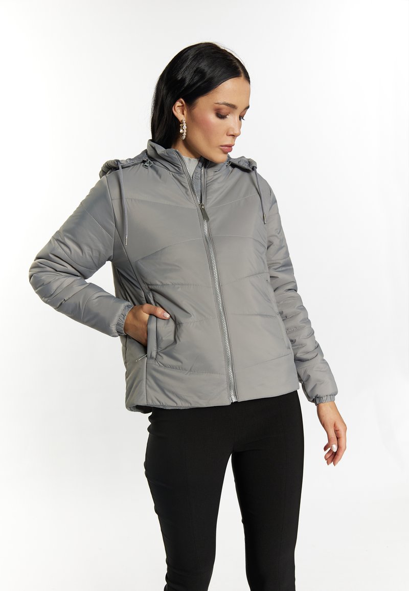 Giacca puffer leggera grigia, con zip e cappuccio, polsini elasticizzati, tasche laterali e dettagli di cucitura orizzontale. Indossata sopra pantaloni neri.