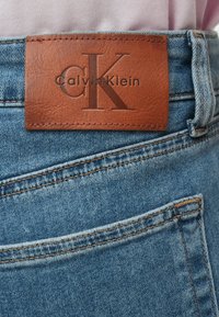 Τα τζιν της Calvin Klein έχουν ανοιχτό μπλε χρώμα, καφέ δερμάτινο λογότυπο με ανάγλυφο σχέδιο και αντίθετες ραφές σε ταμπά χρώμα.