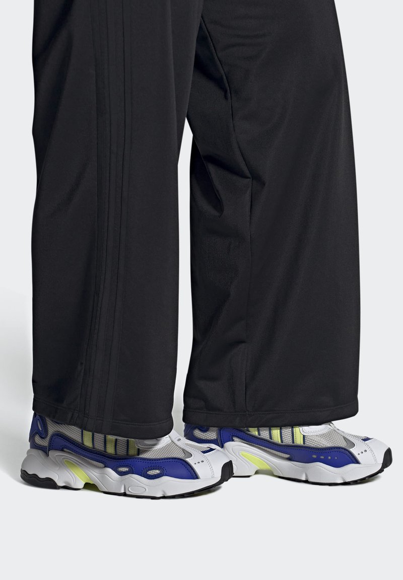 Pantalons de sport noirs avec trois rayures verticales, associés à des baskets blanches imposantes avec des accents bleus, verts et gris.