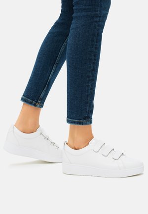 Vita sneakers med kardborreband, ovandel i slätt läder och en tjock vit gummisula. Bärs med blå denimjeans, något upprullade.
