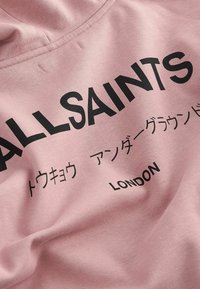 Ružičasta pamučna majica s crnim tekstom. Sadrži "ALLSAINTS" u podebljanom fontu, uz japanske znakove i "LONDON" na dnu. Meka tekstura.