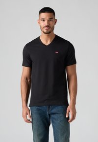 Fekete, pamut anyagú V-nyakú póló, kis piros logóval a mellkason. Kék farmerrel viselve, semleges háttér előtt állva.