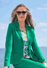 Grüne maßgeschneiderte Jacke und Hose, getragen über einem blumengemusterten Oberteil. Dunkle Sonnenbrille und Creolen vervollständigen den Look.