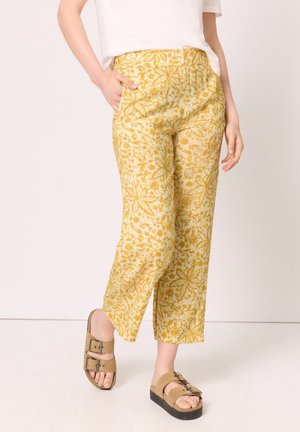 Persona che indossa pantaloni corti gialli a fantasia floreale con tasche laterali e sandali marroni con fibbia su sfondo semplice.