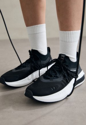 Schwarze Sportschuhe aus strukturiertem Stoff, mit weißer Sohle und schwarzen Schnürsenkeln. Tragen mit weißen gerippten Socken, stehen auf einer hellen Fläche.