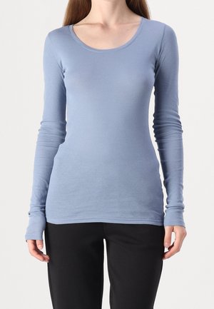 Kobieta w dopasowanej, jasnoniebieskiej koszuli z długim rękawem i okrągłym dekoltem oraz czarnych spodniach, stojąca na jednolitym białym tle.