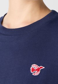 Marineblauw katoenen T-shirt met een geribde ronde hals. Heeft een klein rood geborduurd vogeltje aan de linkerkant. Soepele textuur.