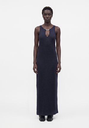 Femme portant une longue robe sans manches bleu marine avec un décolleté en trou de serrure et des chaussures noires, debout devant un fond blanc uni.