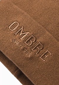 Ruskea neulepipo, jossa on sanan "OMBRE" kirjailu tummemmassa sävyssä. Pipo on pehmeä ja siinä on käännetty reuna.
