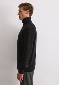 Pull en col roulé noir en tissu doux, présentant un design ajusté, des poignets côtelés et une texture lisse. Porté avec un pantalon gris texturé.