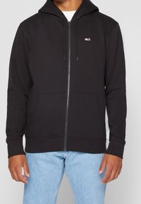 Sudadera negra con cremallera hecha de tejido suave, con un bolsillo frontal, capucha con cordón y un pequeño parche con logo en el pecho.