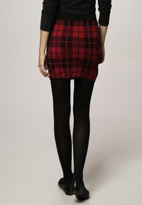 Mini-jupe à carreaux rouges et noirs avec une taille noire ajustée, associée à des collants noirs et des chaussures plates, vue de dos.