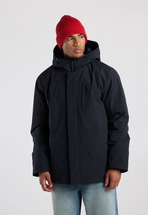 Man met een rode gebreide muts, een zwarte winterjas met kap en lichtblauwe spijkerbroek, kijkt naar links tegen een effen achtergrond.