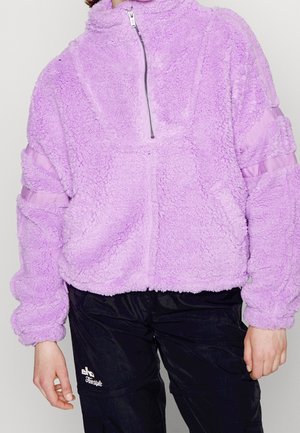 Personne portant une veste polaire lavande à col montant, demi-zip, moelleuse avec des manches froncées et un pantalon noir avec un logo blanc sur la cuisse.