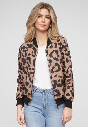 Ung kvinde med blondt hår iført en beige og sort bomberjakke med leopardprint over en hvid top og lyseblå jeans.