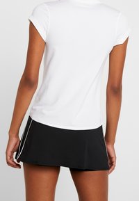 Kobieta w białej bluzce z krótkim rękawem i czarnej spódnicy sportowej z białymi wykończeniami, pokazana od tyłu na jednolitym tle.