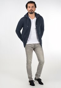 Felpa navy con zip, cappuccio con coulisse, maniche lunghe e due tasche, abbinata a una t-shirt bianca semplice, jeans grigi e sneaker nere.